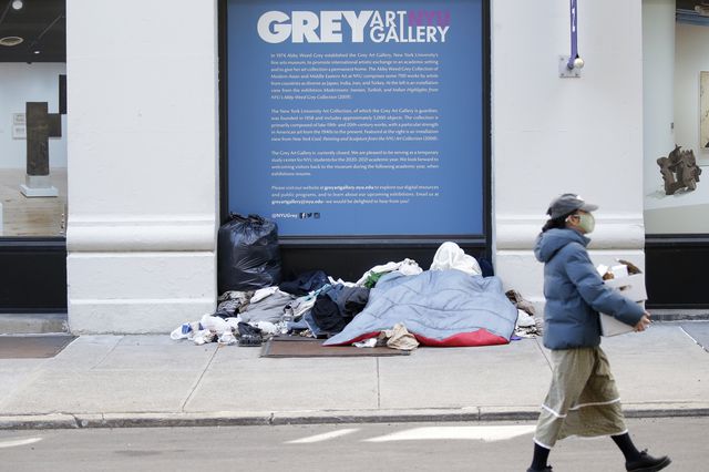 A woman walks by a window for NYU Grey Art Gallery where at least one homeless person has set up blankets and possessions.